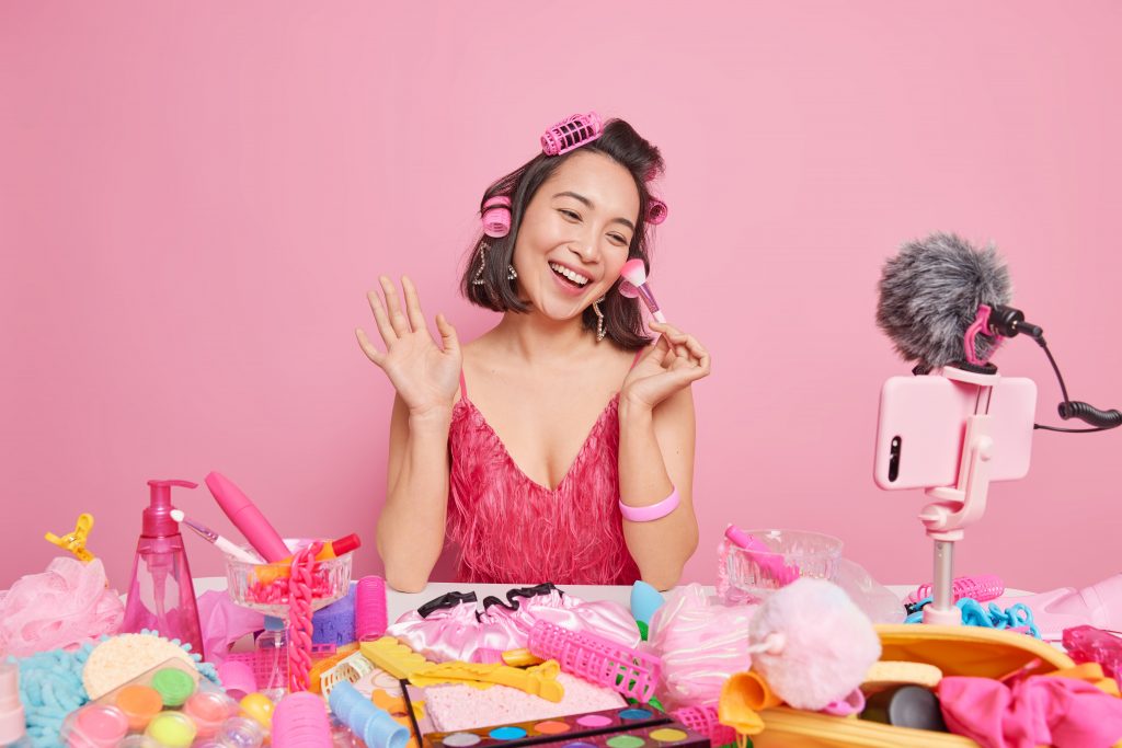female-video-blogger-streaming-online-workshop-uses-cosmetic-brush-makes-hairstyle-with-rollers-sits-front-smartphone-webcam-wears-fashionable-pink-dress
