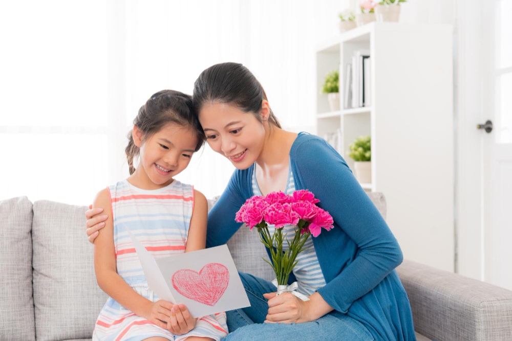 asian-chinese-woman-holding-pink-bouquet-with-cute-little-girl-sitting-together-living-room-sofa-together-looking-card-content-gift-from-family-mother-s-day-with-copyspace-min
